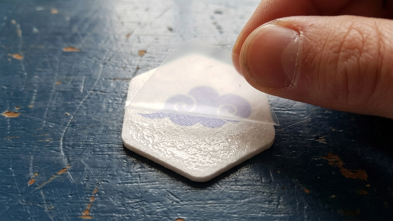 A hand peeling the clear liner from the adhesive back of a hexagonal cloud-marked patch.